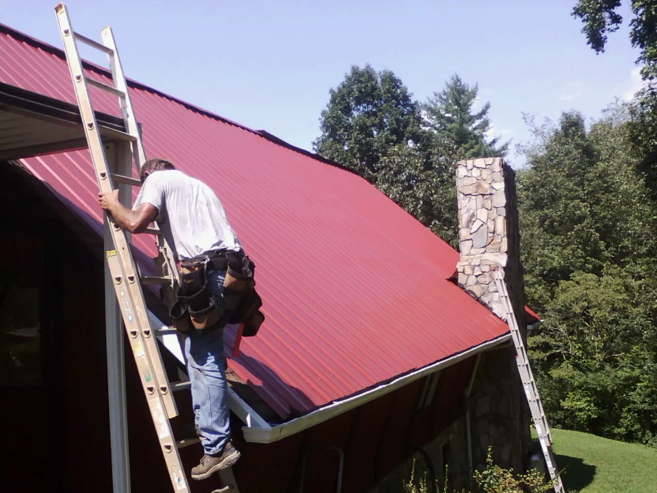 Roofer on a ladder beside a red metal roof and stone chimney — Soffit & Fascia Repair by Roofing Brothers in Idaho City